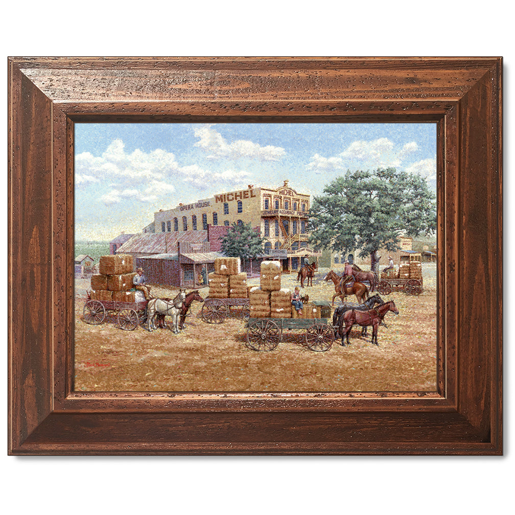 canvas print in a brown wood frame depicting a main street in a 
western town with cotton wagons hauling large cotton bails