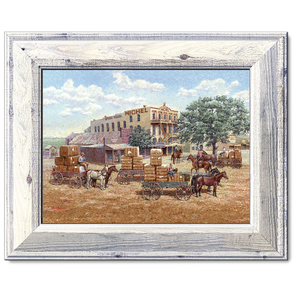 canvas print in a white wood frame depicting a main street in a 
western town with cotton wagons hauling large cotton bails